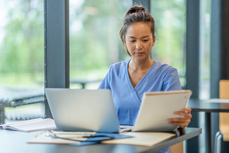 Young female pretty nurse taking SimpleNursing NCLEX practice text at a desk with a laptop and a notebook.