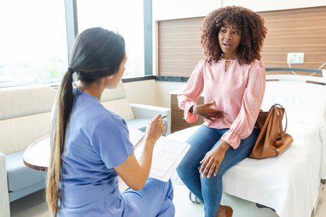 A young Black woman patient touches her hand to her stomach while on an examination bed as a young woman nurse in scrubs examines her.
