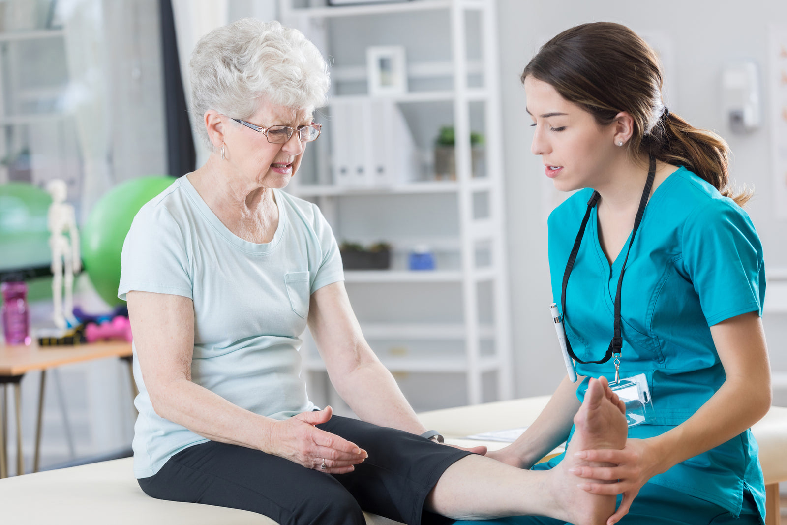 Young female nurse tests the flexion of an older lady's ankle and foot for injury and range of motion in a hospital examination room.