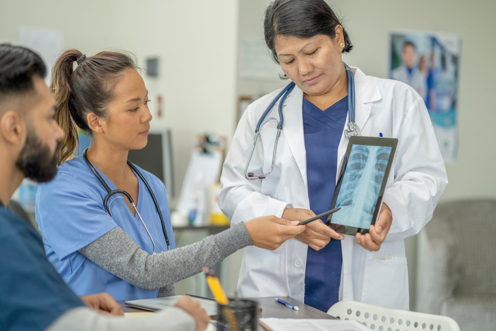 A female nursing school teacher in a white lab coat holds a tablet and shows two young nursing students a chest x-ray of ribs.