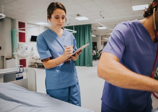 A young brunette nurse takes notes onto a chart with clipboard next to a male nurse with stethoscope