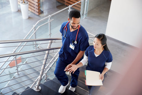 Two young nurses wearing navy blue scrubs walk up a stairwell stairs together nurses working together