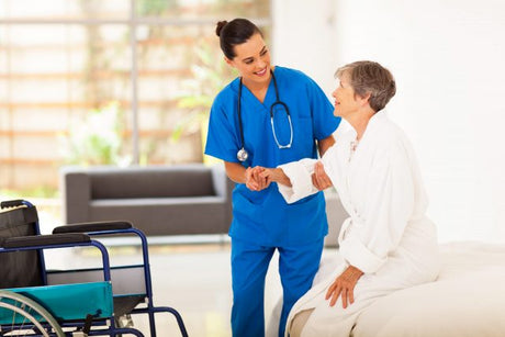A young female nurse wearing blue scrubs and a stethoscope helps an older woman out of bed and into a wheelchair dvt nursing intervention patient