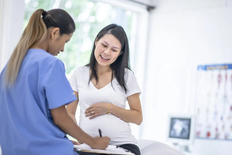 A pregnant Asian woman sits with her hands on her belly as she visits a mixed race female doctor. The doctor is wearing blue scrubs and has her back to the camera.