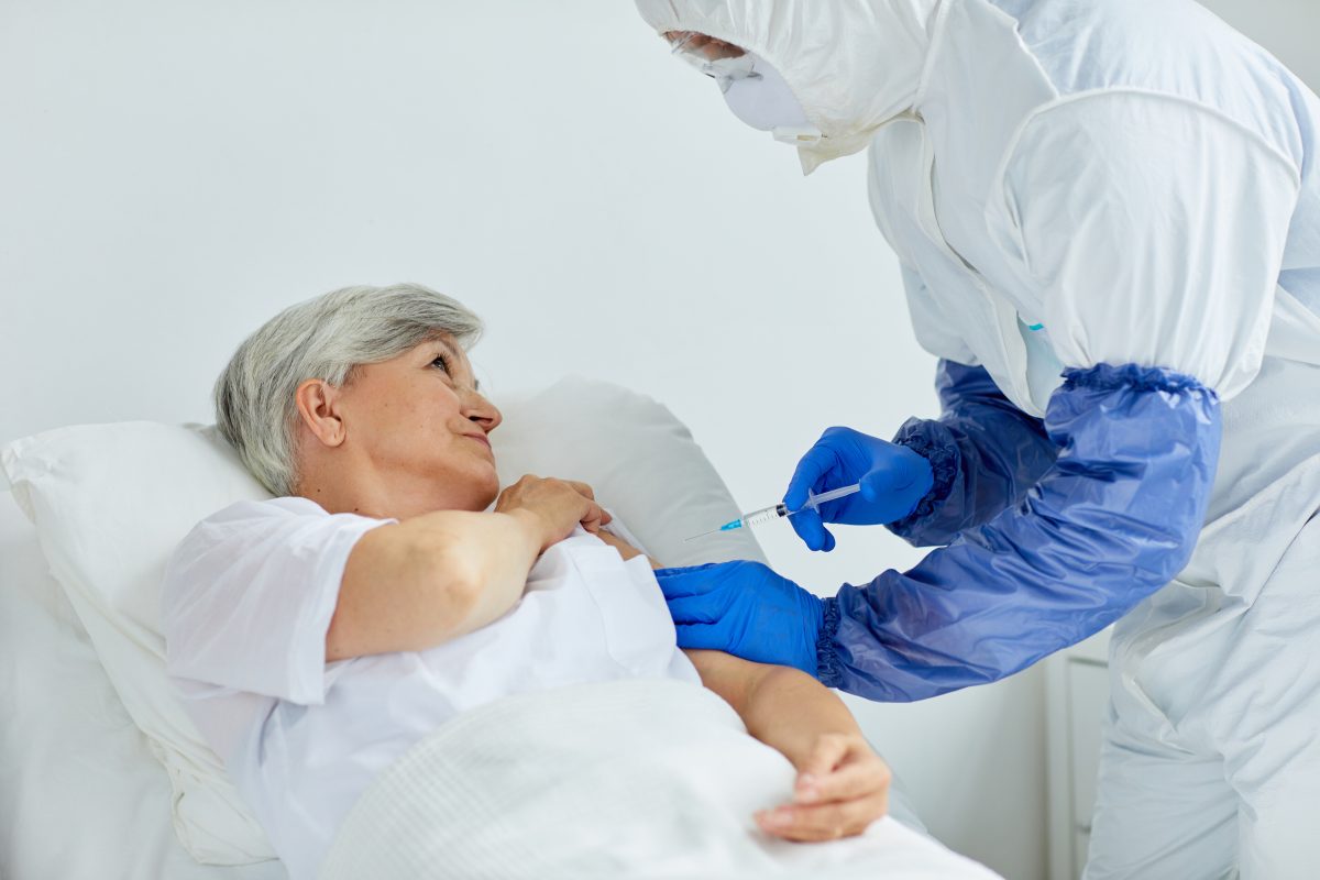 Modern nurse wearing protective suit giving injection to mature woman lying on bed in hospital ward. Treating Senior Patient With Injection.