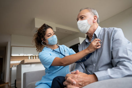 Nurse doing a medical exam on a senior patient at home during the COVID-19 pandemic and wearing facemasks â healthcare and medicine concepts