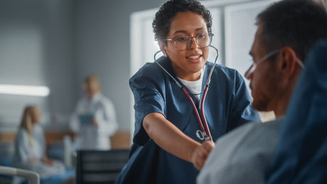 Hospital Ward: Friendly Black Head Nurse Uses Stethoscope to Listen to Heartbeat and Lungs of Recovering Male Patient Resting in Bed, Does Checkup. Man Getting well after Successful Surgery