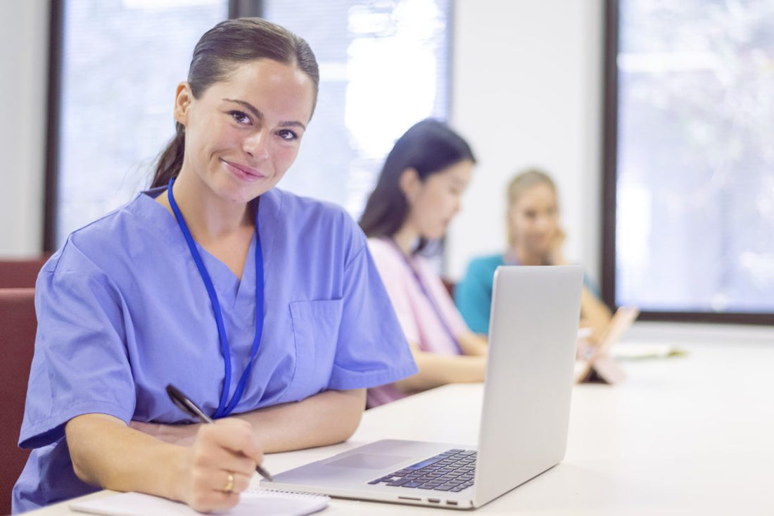 A row of female university nursing students attending a healthcare lecture in a modern classroom setting. Shot focused on a Caucasian trainee nurse who is smiling and looking directly at the camera. All students are wearing medical scrubs.