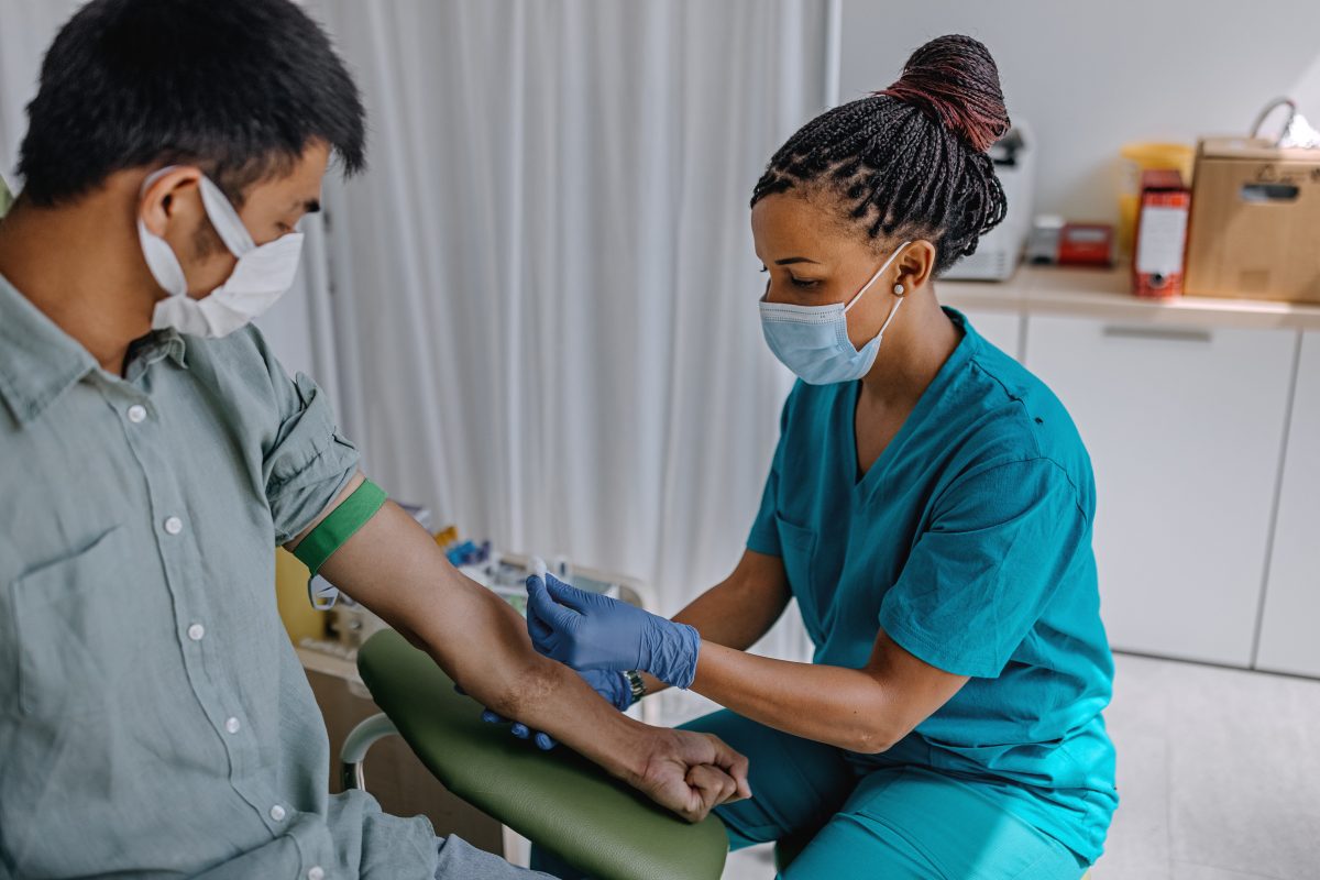 African American Black female nurse in a mask drawing blood from a masked man .How to Become a Phlebotomist + Salary Expectations