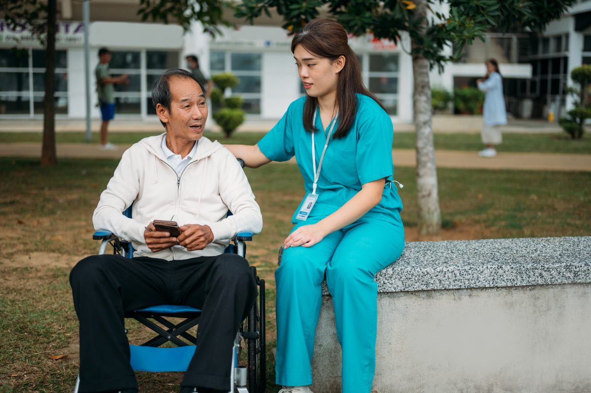 A young Asian woman nurse sits with an older Asian man in a wheelchair in a nursing home hospital hospice facility.
