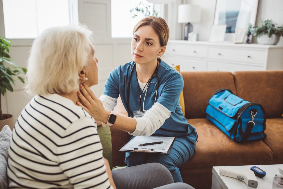 Nurse visiting senior patient at home. She is talking with senior woman about her condition and checking thyroid gland.