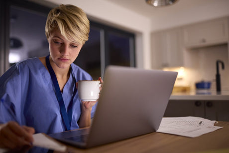 Tired Woman Wearing Medical Scrubs Working Or Studying On Laptop At Home At Night