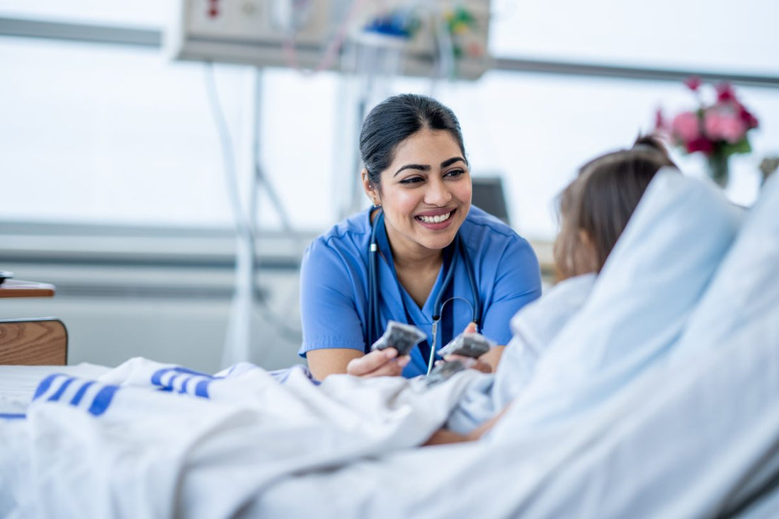 A female nurse of Middle Eastern decent sits at the edge of a hospital bed as she check in on her young patient. She is wearing blue scrubs and is attempting to cheer the young girl up. Nurse Checking on a Patient