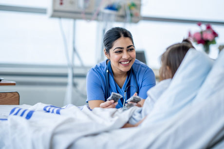 A female nurse of Middle Eastern decent sits at the edge of a hospital bed as she check in on her young patient. She is wearing blue scrubs and is attempting to cheer the young girl up. Nurse Checking on a Patient