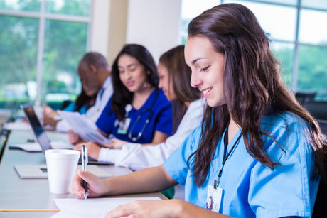 Young adult Hispanic nursing student is taking notes for her nursing exit exams. She is wearing blue scrubs and a student ID badge.