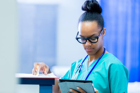 Young adult African American female nurse or doctor in hospital emergency room is checking paper charts and digital charts in modern digital tablet. Woman is wearing scrubs and glasses.