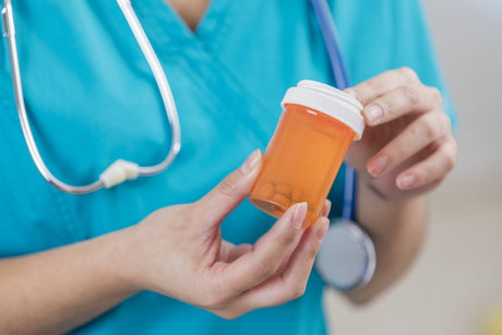 Nurse holds a prescription bottle containing medication tablets. The nurse is wearing scrubs and a stethoscope.