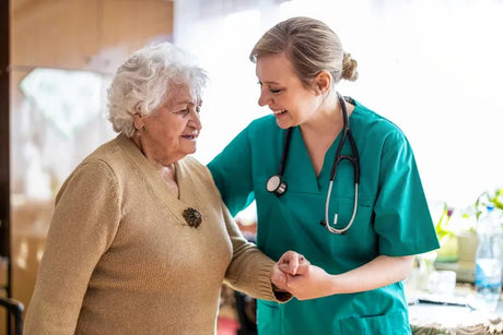A female nurse in scrubs and a stethoscope assists and aids an older woman following maslow's hierarchy in nursing header